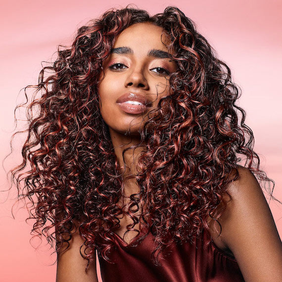 Model with long, dark, glossy, curly hair featuring coral highlights.