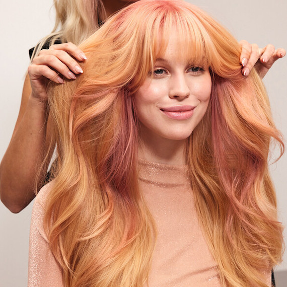 A hairdresser styles a model’s long, rose gold, wavy hair.