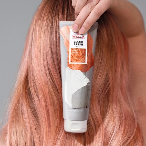 Model with pinky blonde hair holds up a bottle of the Color Fresh Mask in Peach Blush.