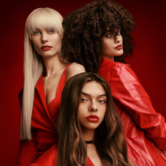 3 models all wearing red stand in front of a red backdrop. One has blonde hair, one has dark curly hair & one has brown hair