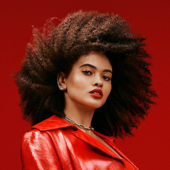 Headshot of a model in front of a red backdrop wearing a red jacket and lipstick. They have voluminous brown curly hair 