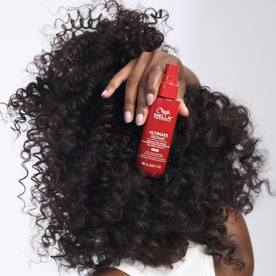 : A model's face is concealed by their curly brown hair. They hold a bottle of Miracle Hair Rescue Treatment over their head