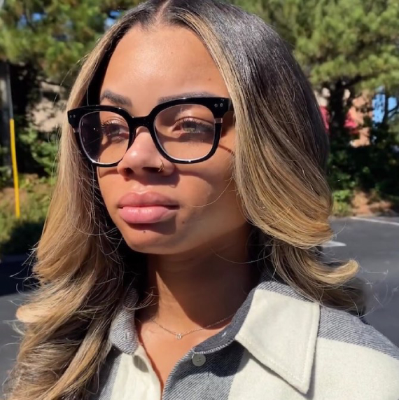 Headshot of a person with blonde balayage highlights on brown hair. They wear black rimmed glasses and a gray/white shirt