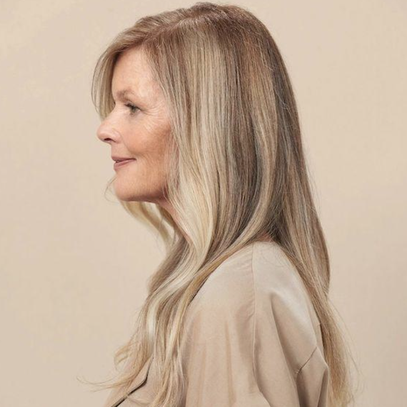 Side profile of a person with long blonde balayage hair. They wear beige clothing and stand in front of a beige backdrop