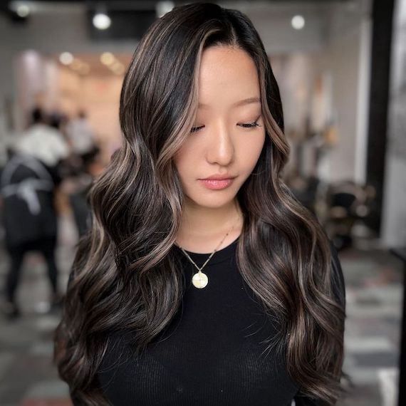 Model with long, wavy, black hair with ashy highlights glances downwards.