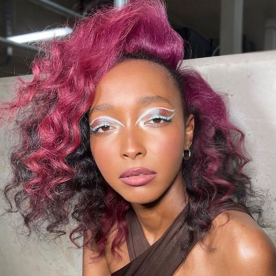 Model with long, curly, black hair and magenta color block highlights looks directly into the camera.