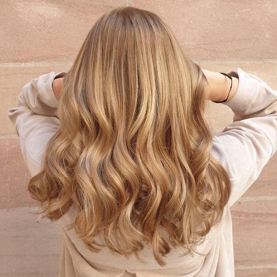 A person with long, wavy honey blonde hair stood in front of a beige brick wall with their back facing the camera.