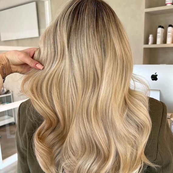A person with long, honey blonde babylights sits in a salon with her back to the camera to show off her newly colored hair