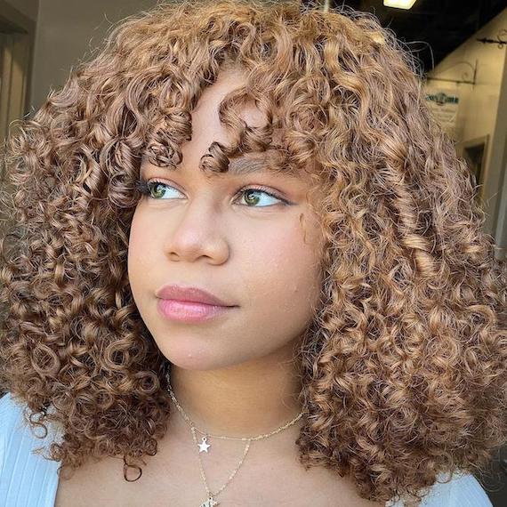 Headshot of a person with deep honey blonde chunky highlights on curly hair and hazel green eyes