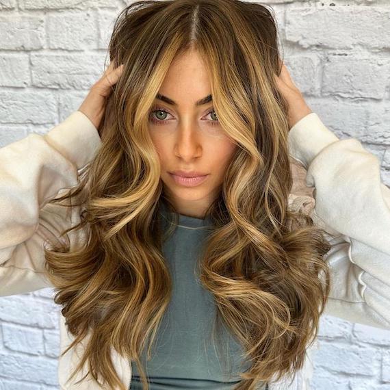 A person with bronde hair and honey highlights wears a white sweater and stands in front of a white brick wall