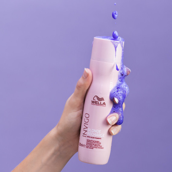 Close-up of a hand holding a bottle of Wella Professionals Invigo Blonde Recharge Shampoo in front of a purple backdrop