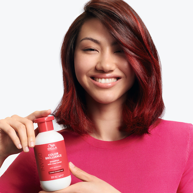 Model with dark red, shoulder-length hair smiles at the camera while holding a bottle of INVIGO Color Brilliance Color Protection Shampoo.