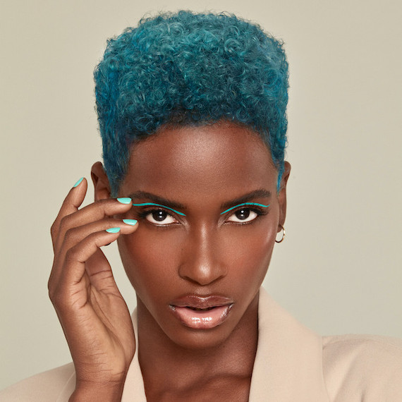 Model with short, coily, teal hair hair, green eyeshadow and green nail polish faces the camera.