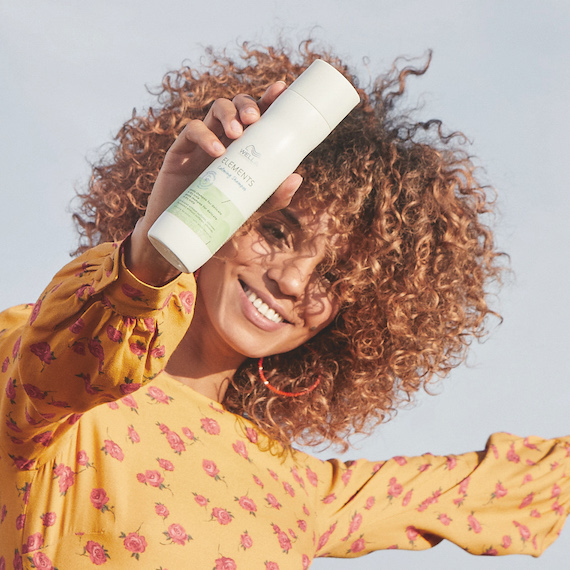 Model with curly, brown hair holds up a bottle of Elements Shampoo.