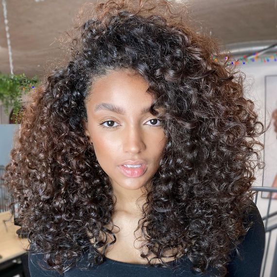 Model with dark brown, curly hair and bronze highlights faces camera.