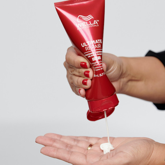 A red bottle of Ultimate Repair Conditioner being squeezed into a palm. The person's nails are painted red to match