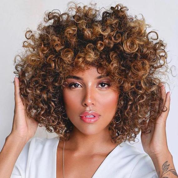 Woman looking into the camera with beautiful, curly, brown hair and caramel highlights.