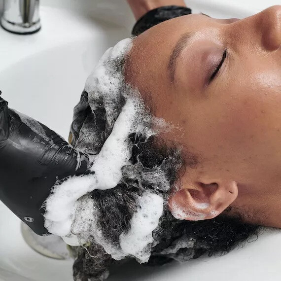 A model’s dark, curly hair is washed at the sink by a hairdresser.