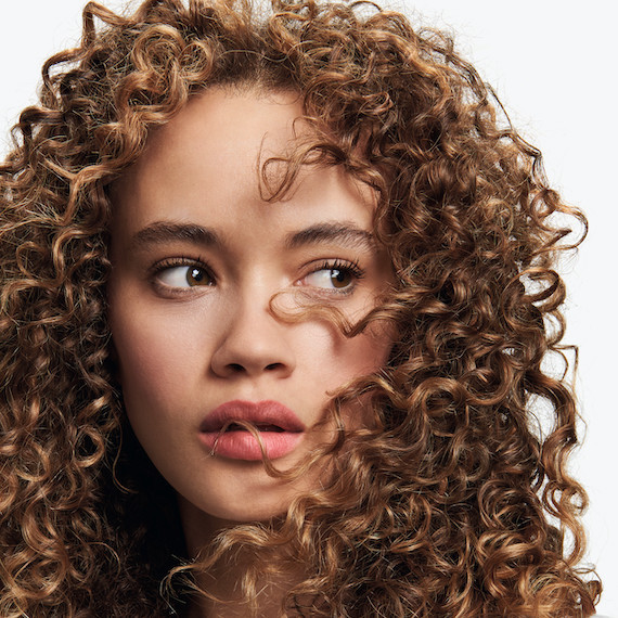 Model with shiny, curly, brown hair looks off to the side.