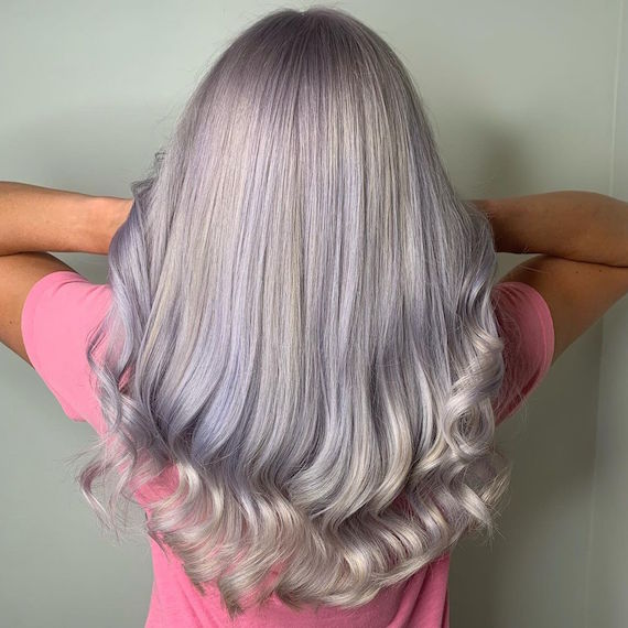 A person wearing a pink t-shirt faces away from the camera to reveal their full head of silver lilac hair