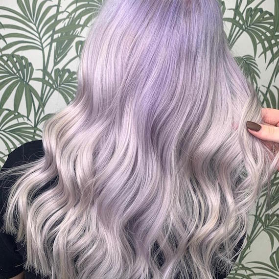 A person with wavy lilac ombre hair faces a wall decorated with green leaf printed wallpaper 