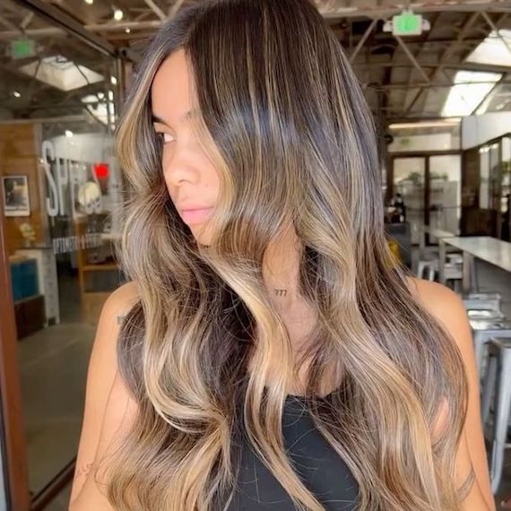 Model with dark roots and golden highlights in their long wavy hair looks over their shoulder