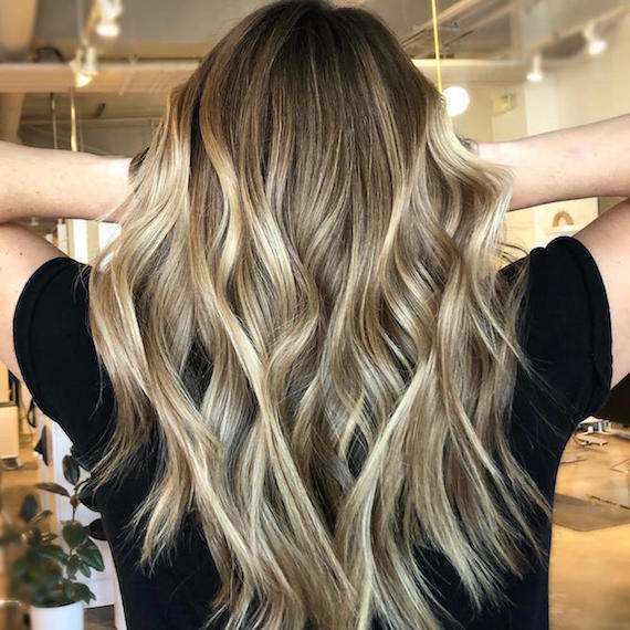 Hinterkopf einer Frau mit langem, gewelltem Haar und blonder Balayage, kreiert mit Wella Professionals. 