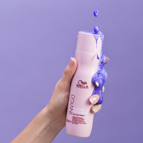 Hand holding a bottle of INVIGO Cool Blonde Shampoo, with purple shampoo spilling down the sides. 