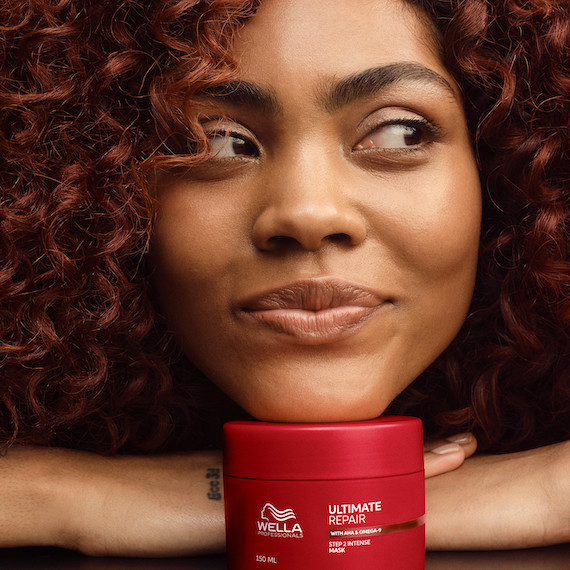 Model with red and dark brown curly hair rests chin on tub of ULTIMATE REPAIR Mask.