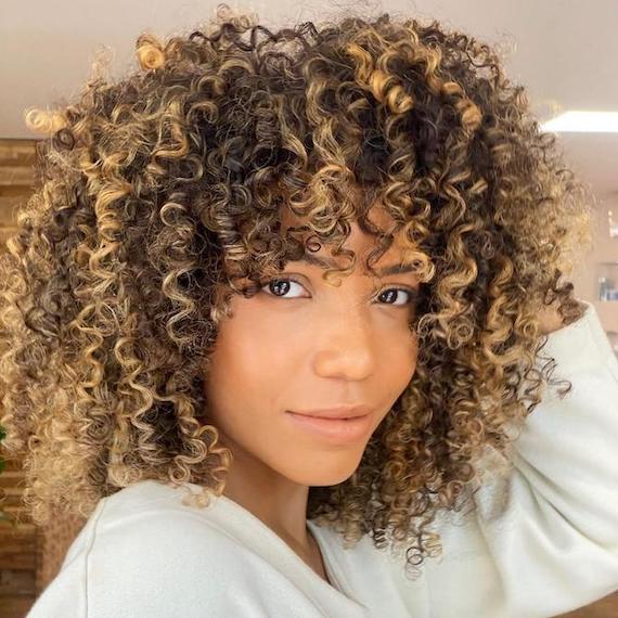 Woman smiling at camera with brunette curls and caramel blonde highlights, created using Wella Professionals. 