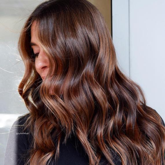 A person with long, wavy mocha brown hair looks to the side so that their hair conceals most of their face