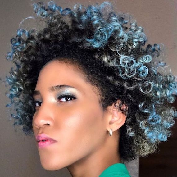 Model smiles at camera with dark brown curly hair and pastel blue balayage.