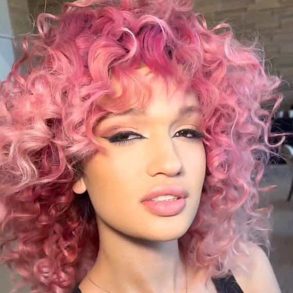 Model with thick, curly, pink ombre hair looks into the camera.