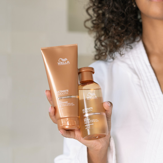 Model with dark brown, curly hair holding bottles of ULTIMATE SMOOTH Shampoo and ULTIMATE SMOOTH Conditioner.