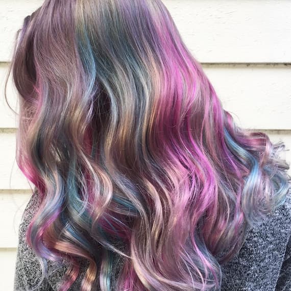 Image of the back of a woman’s head, showing purple and blue unicorn-inspired rainbow hair styled in loose waves. Look created by Wella Professionals.