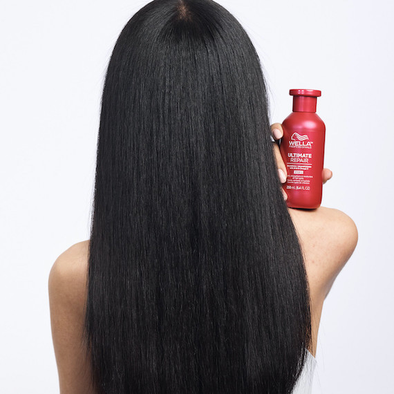 A model with long dark hair faces away from the camera holding a bottle of Ultimate Repair Shampoo on their shoulder