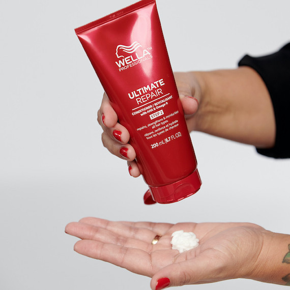 Close-up of a red bottle of Ultimate Repair Conditioner being squeezed into someone's palm. They have red painted nails