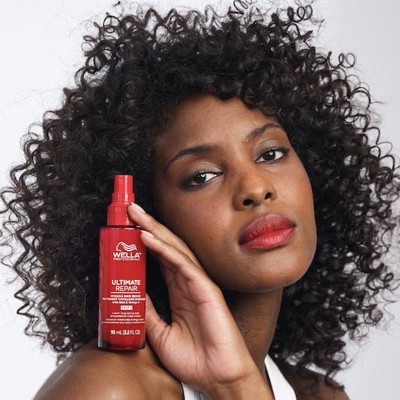 Ein Model mit lockigem dunklem Haar und rotem Lippenstift hält eine Flasche Ultimate Repair Miracle Hair Rescue Treatment auf der Schulter.