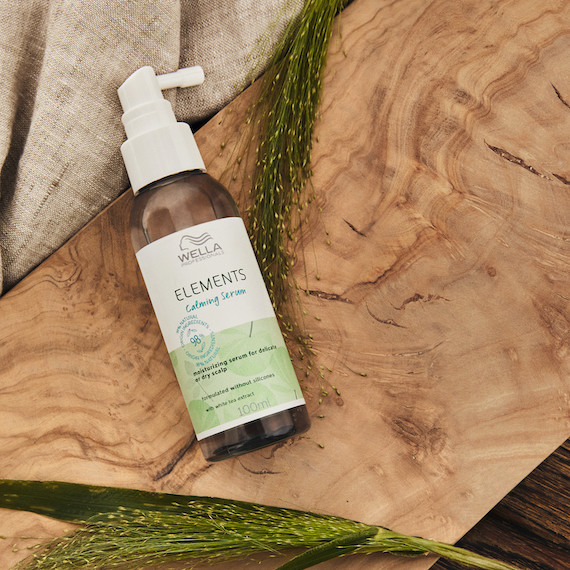A bottle of Elements Calming Serum pictured against a wooden board, beige fabric and green plants