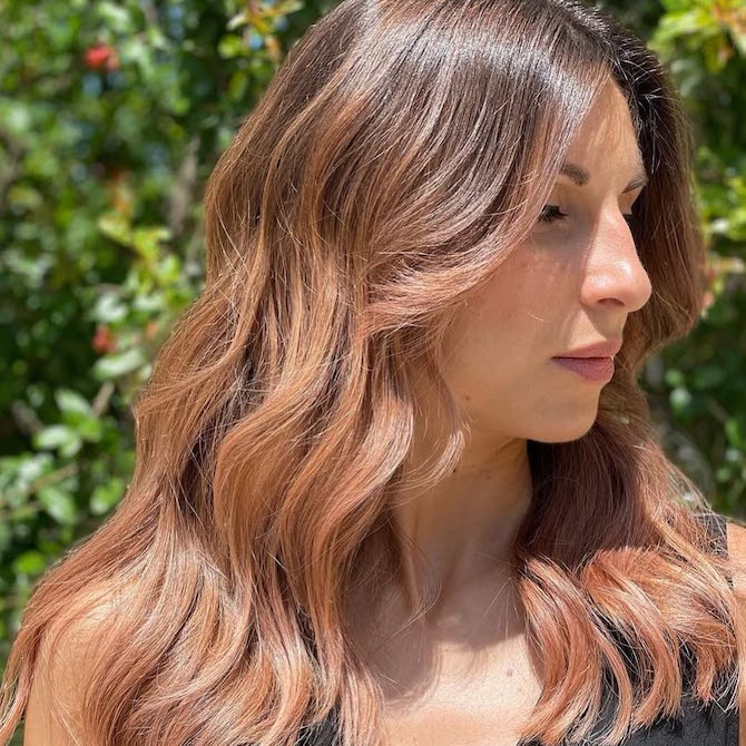 Model with long, medium brown hair featuring a rose gold balayage through mid-lengths and ends.