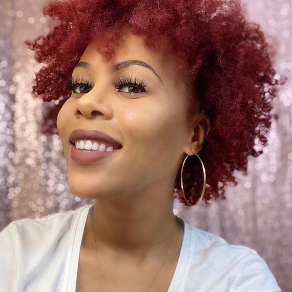 Headshot of a person with short, curly ruby red and purple hair. They wear a white top and gold hoop earrings