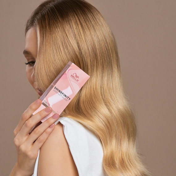 Ein Model mit langem blondem Haar blickt zur Seite, während es eine Packung Shinefinity Color Glaze über die Schulter hält