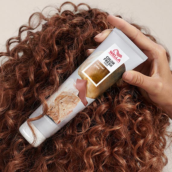 A model with brown, curly hair holds up a bottle of the Color Fresh Mask in Caramel Glaze.