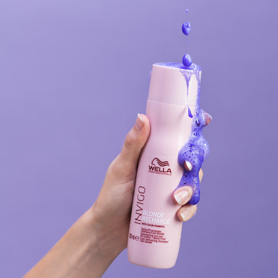 A hand holds Wella’s INVIGO Cool Blonde Shampoo on a purple background. The shampoo drips down the side of the bottle.