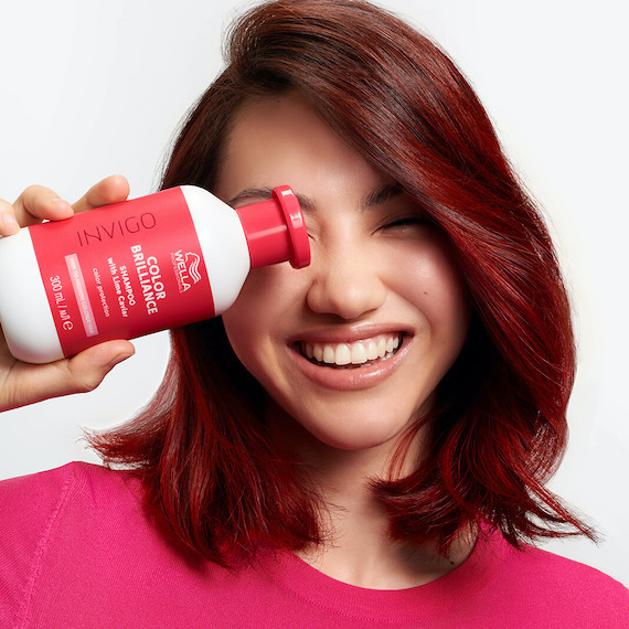 Model with dark brown hair and red highlights holds up a bottle of INVIGO Color Brilliance Color Protection Shampoo.