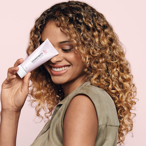 Model with curly, glossy hair faces the camera smiling, while holding a tube of Wella Shinefinity Glaze.
