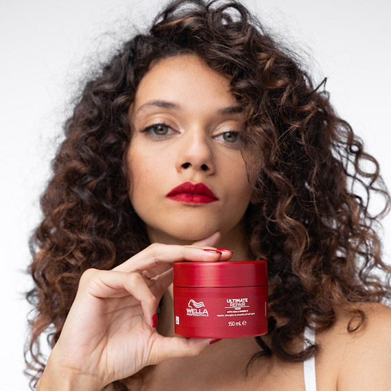 Model with dark brown, curly hair holds a tub of ULTIMATE REPAIR Mask.