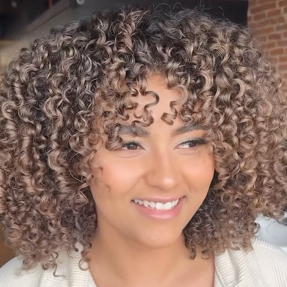 Model with shoulder-length, brown curly hair and bronze highlights.