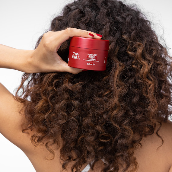 Model with long, dark brown, curly hair holds up a tub of ULTIMATE REPAIR Mask.