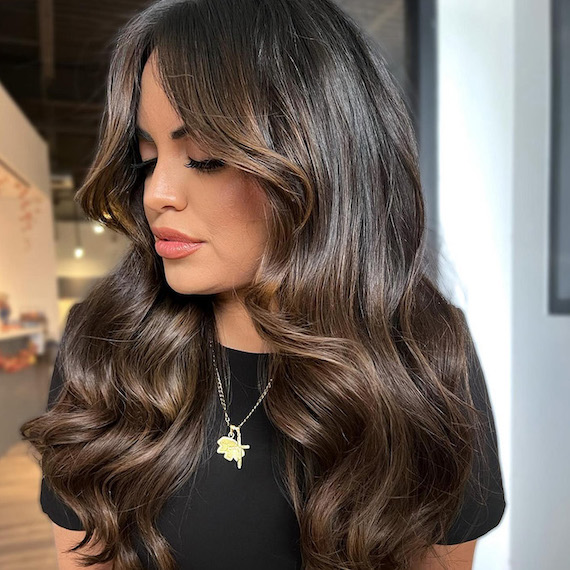 Model with long, loosely curled, dark brown hair featuring dimensional highlights.